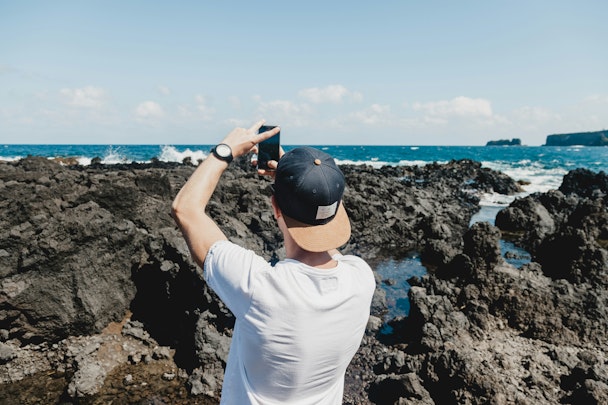 A man uses his phone to take a photo of the sea and a rocky shoreline