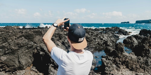 A man uses his phone to take a photo of the sea and a rocky shoreline