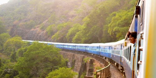 A train carrying a passenger (leaning out of the window) travels through green mountains