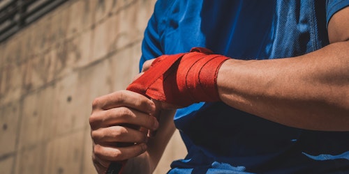 A man in a blue t-shirt wraps red tape around his knuckles as if preparing to box