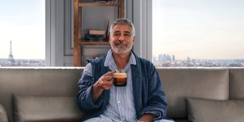 George Clooney sits on a sofa holding a cup of black coffee