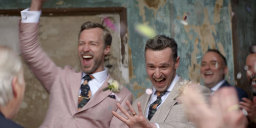 Two men in suits emerging from their wedding ceremony, one with his arm raised in celebration, the other looking at his wedding ring, as guests throw confetti